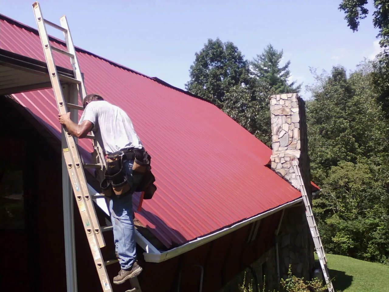 Roofer on a ladder beside a red metal roof and stone chimney — Soffit & Fascia Repair by Roofing Brothers in Bedford Hills